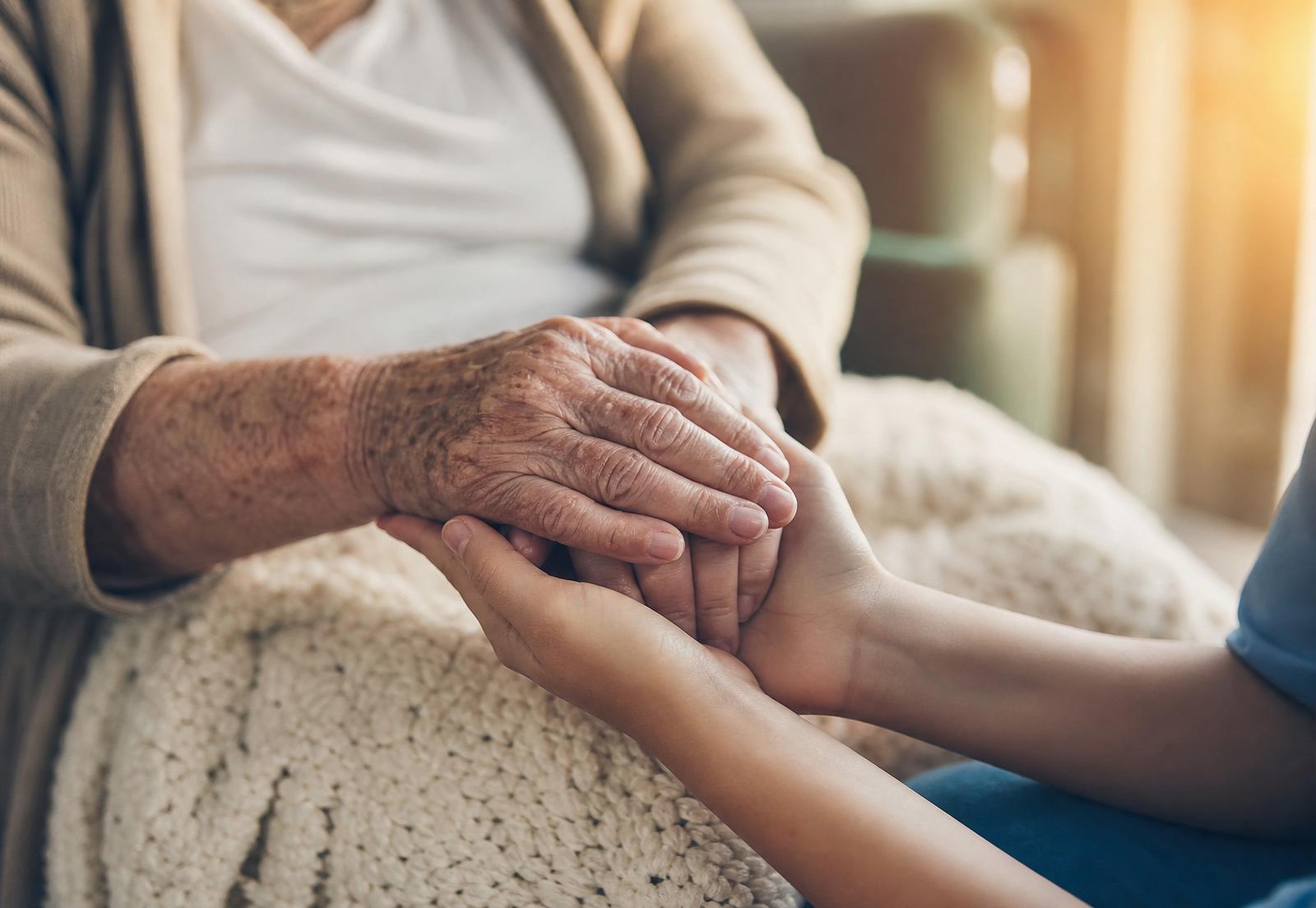 Close-up of an elderly woman's hands held by a caregiver on a soft cream blanket